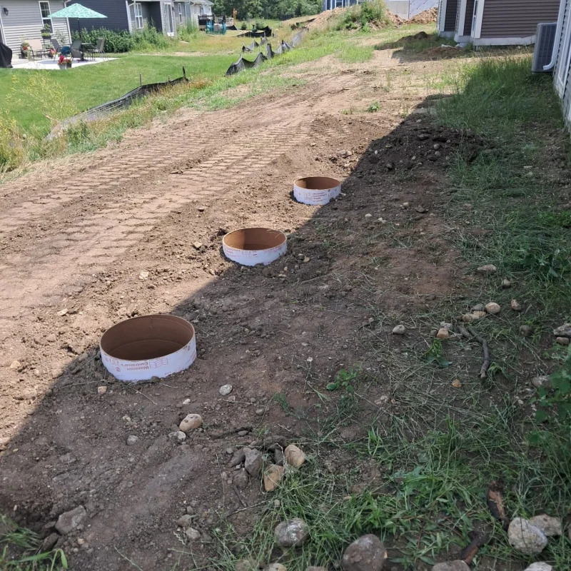 Concrete pier forms set in an excavated strip of ground beside a house, gravel and turf around them
