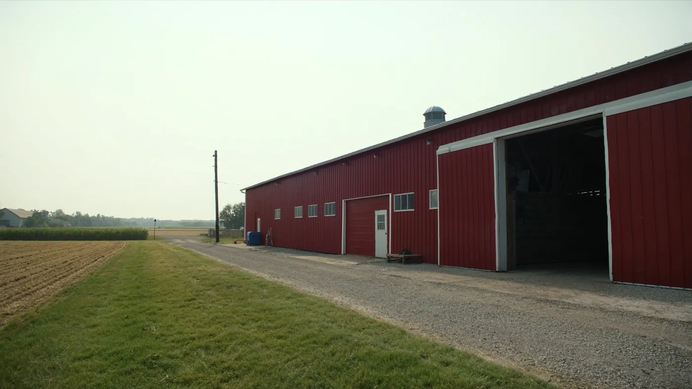 A red steel machine shed with its door open beside a gravel drive and a corn field under a pale morning sky
