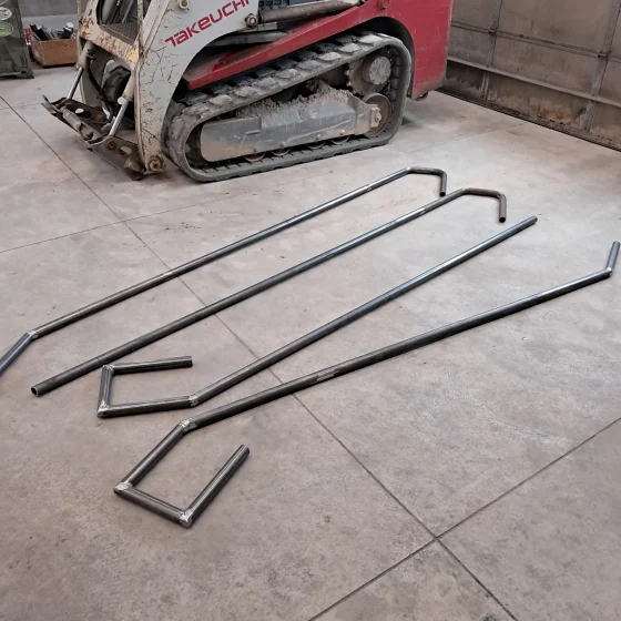 Lengths of steel tube bent into rail sections laid out on a shop floor in front of a tracked skid steer