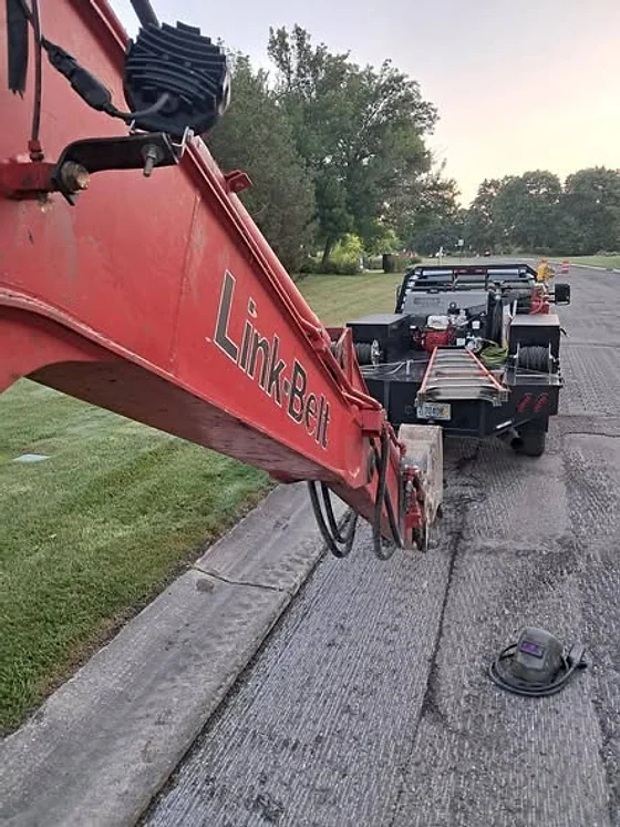 The red boom of a Link-Belt excavator lowered onto a road beside the welding rig