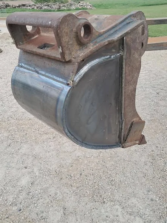 An excavator bucket standing on gravel with new steel plates welded into its sides and floor