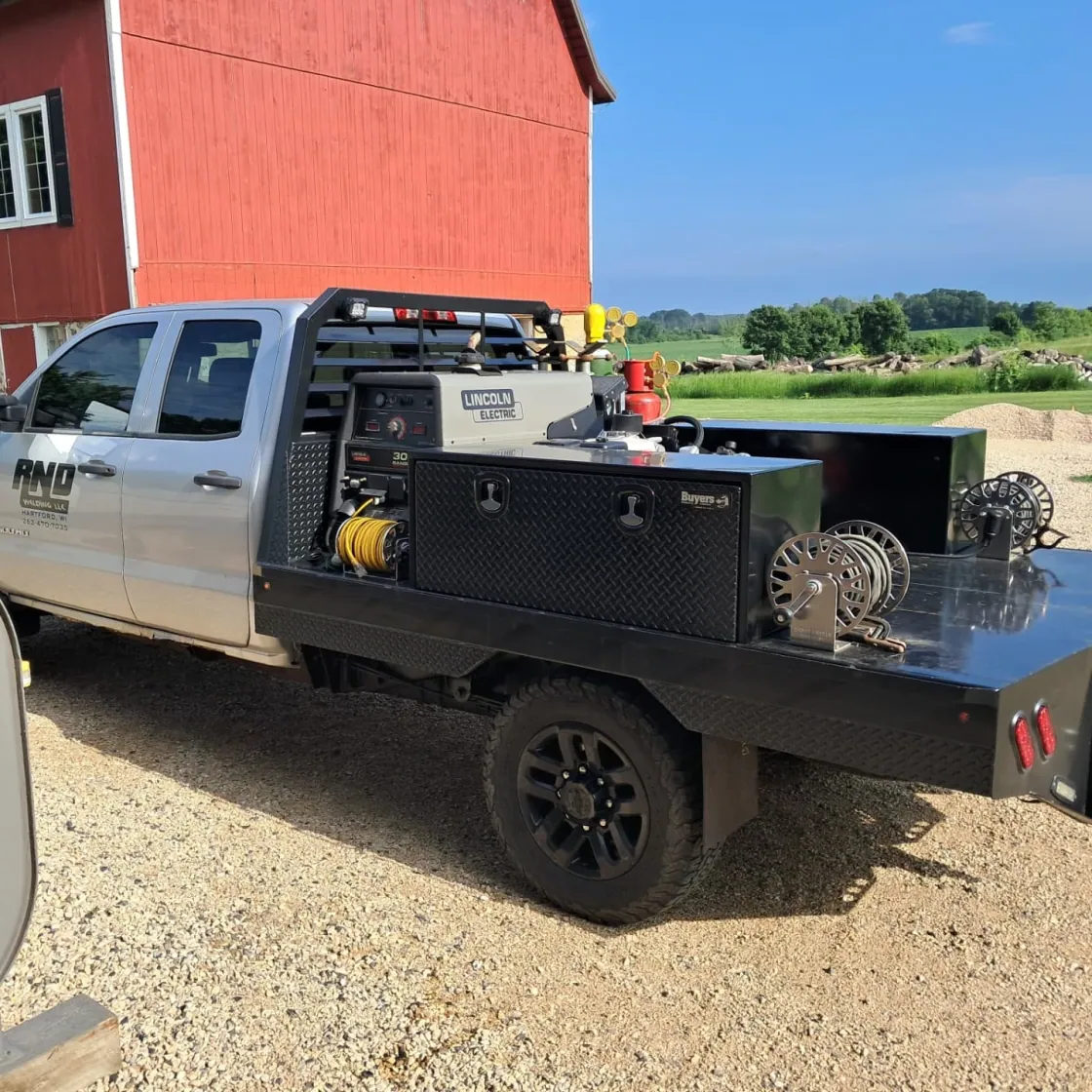 A silver flatbed pickup with RND Welding LLC on the door, an engine driven welder, tool boxes and cable reels on the bed, parked on gravel beside a red barn
