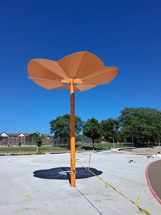 An orange steel shade structure shaped like a flower on a single post at a new playground under a blue sky